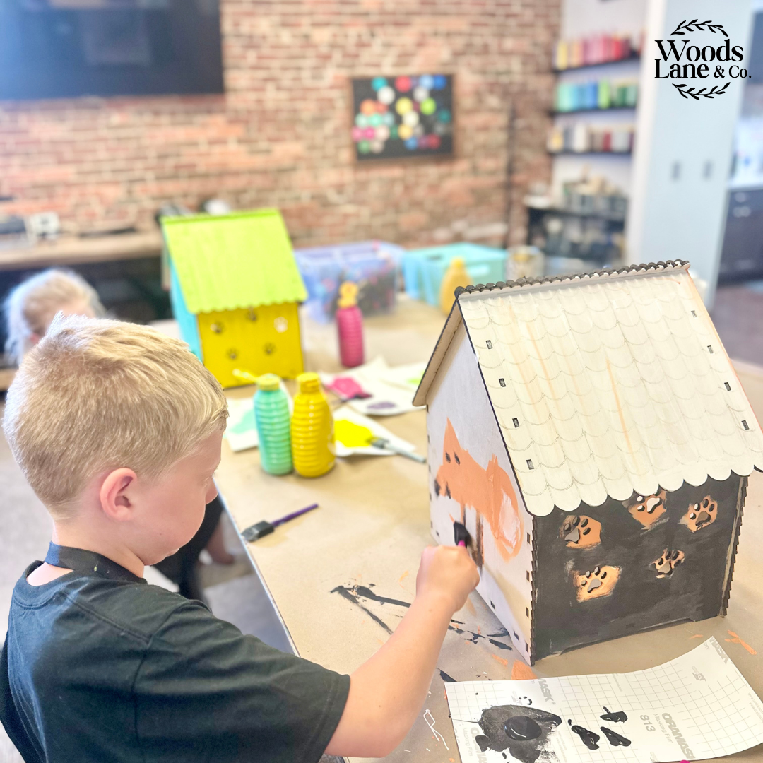 A young boy painting a decorative cardboard house at a craft table in a classroom, with other colorful houses and art supplies visible in the background. The Woods Lane & Co.