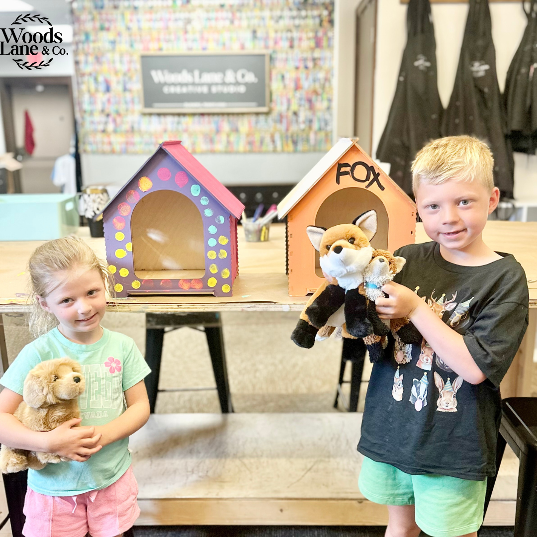 Two children standing in a workshop holding plush toys, with two small decorative stuffy houses on a table behind them. A sign reading Woods Lane & Co.