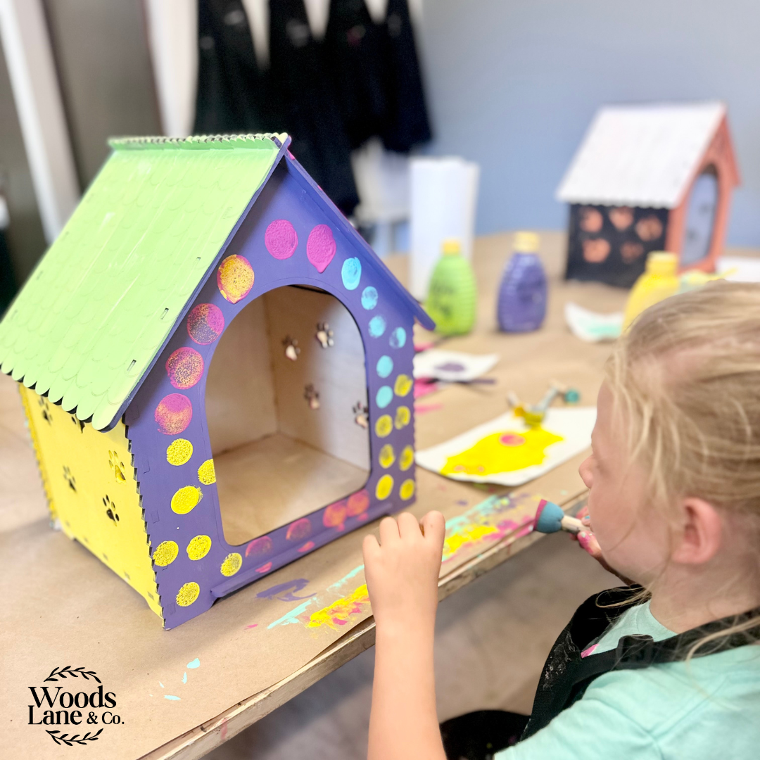 A child painting a colorful wooden house-shaped craft or dog house on a table, with a second finished house in the background. The main house is painted purple, yellow, and green with pink, blue, and yellow polka dots.