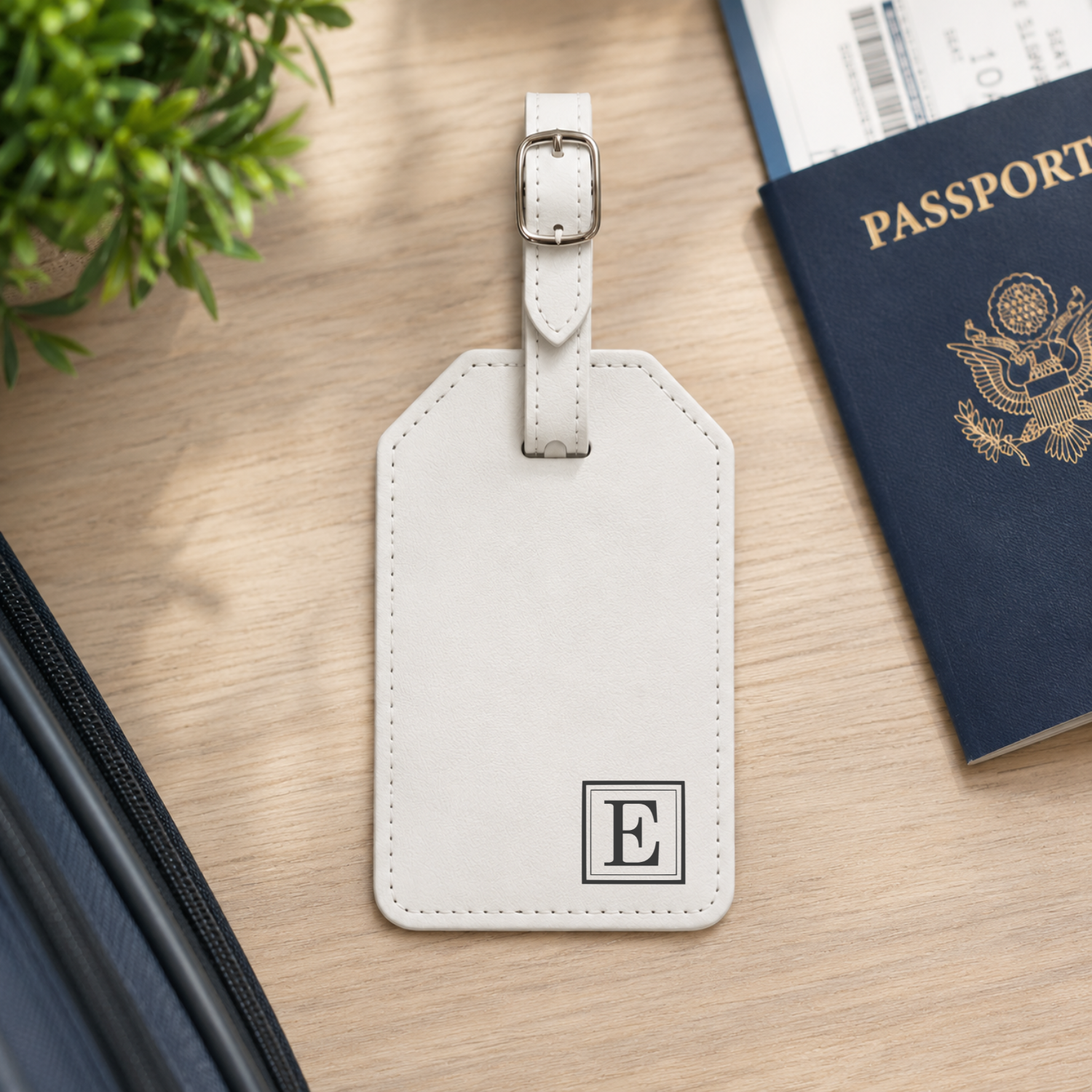 A light beige leather luggage tag with a silver buckle and a monogrammed initial 'E' in the bottom corner, styled on a wooden desk next to a passport and a plant.