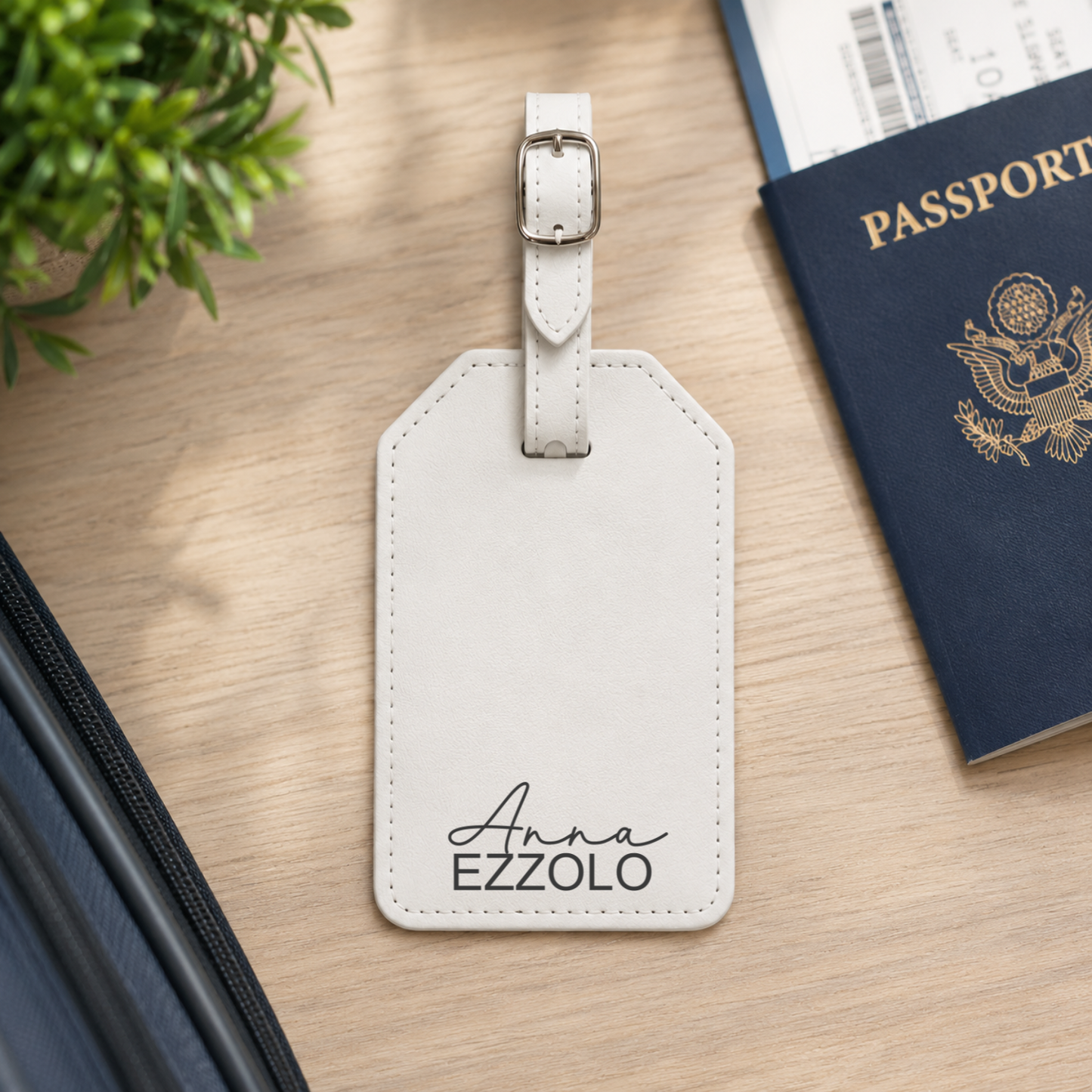 A personalized white leather luggage tag with a silver buckle, featuring the name 'Anna EZZOLO' printed in black script and block lettering. The tag is styled on a light wood desk next to a passport, a zippered case, and a small green plant.