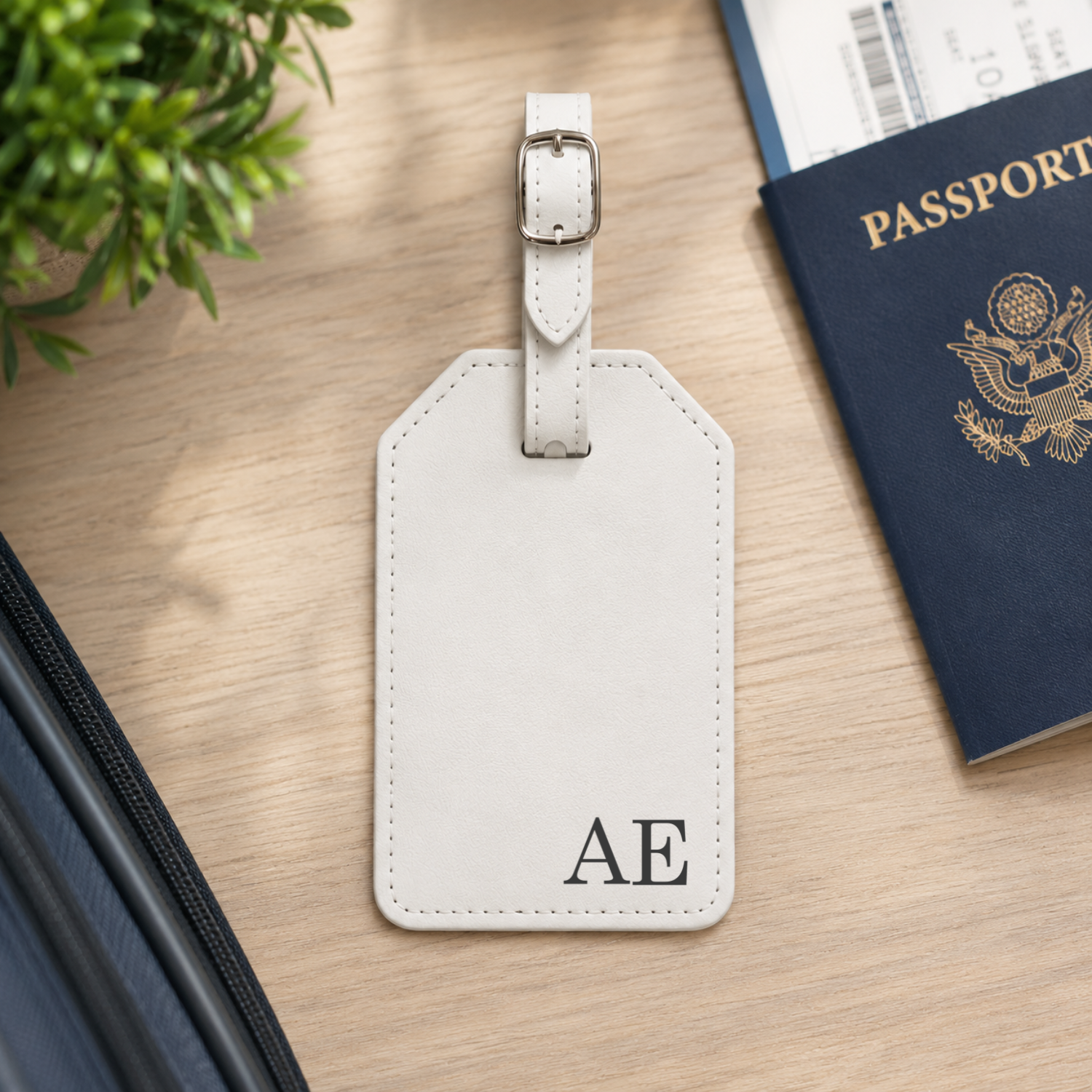 A personalized light beige leather luggage tag with a silver buckle, featuring the monogram 'AE' in black on the bottom right. The tag is styled on a light wood desk next to a navy blue passport, a small green plant, and a black zippered pouch.