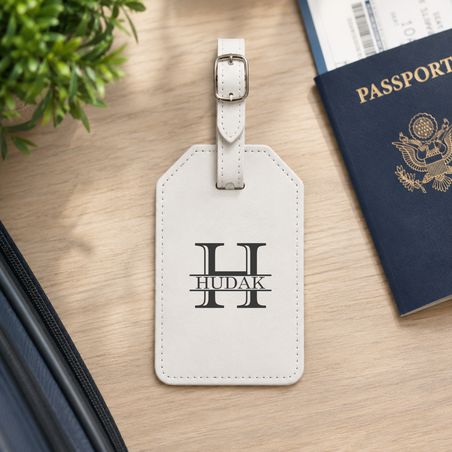 A white leather luggage tag with a silver buckle, personalized with a black monogram 'H' and the name 'HUDAK' in the center, displayed on a wooden desk next to a passport and a small plant.