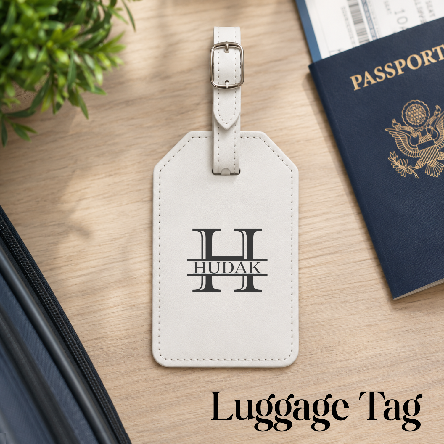A personalized off-white leather luggage tag with a silver buckle, featuring a black monogram 'H' and the name 'HUDAK'. It is displayed on a light wood desk next to a navy blue passport, with the text 'Luggage Tag' at the bottom.